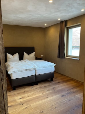 A bedroom with a double bed, wooden flooring, a window with curtains, and a ceiling with recessed lighting.