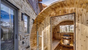 A wooden sauna with a stone exterior, a wooden deck, and a view of snow-capped mountains.
