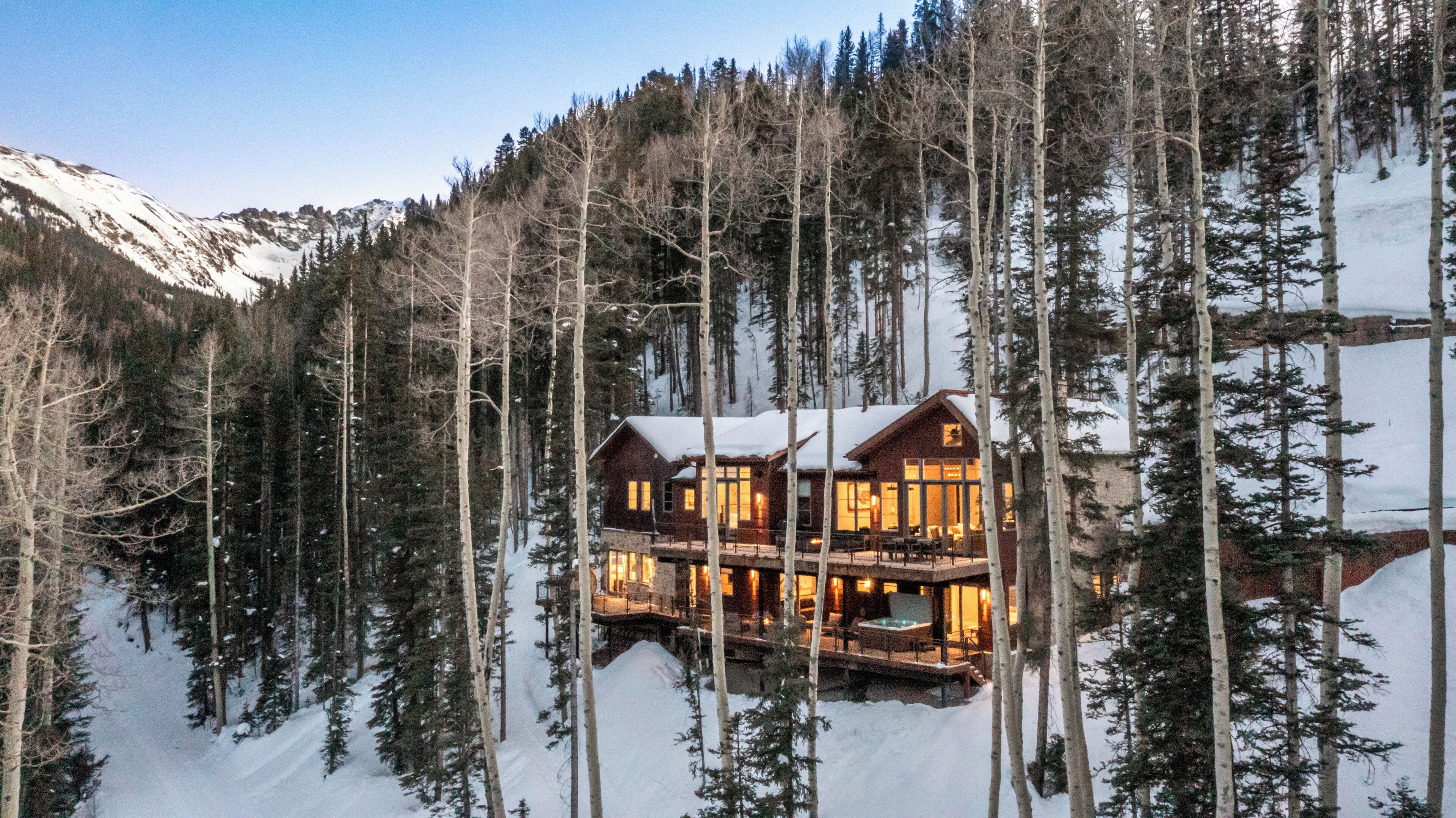 A log cabin with multiple windows and a balcony, surrounded by tall trees and snow-covered ground.