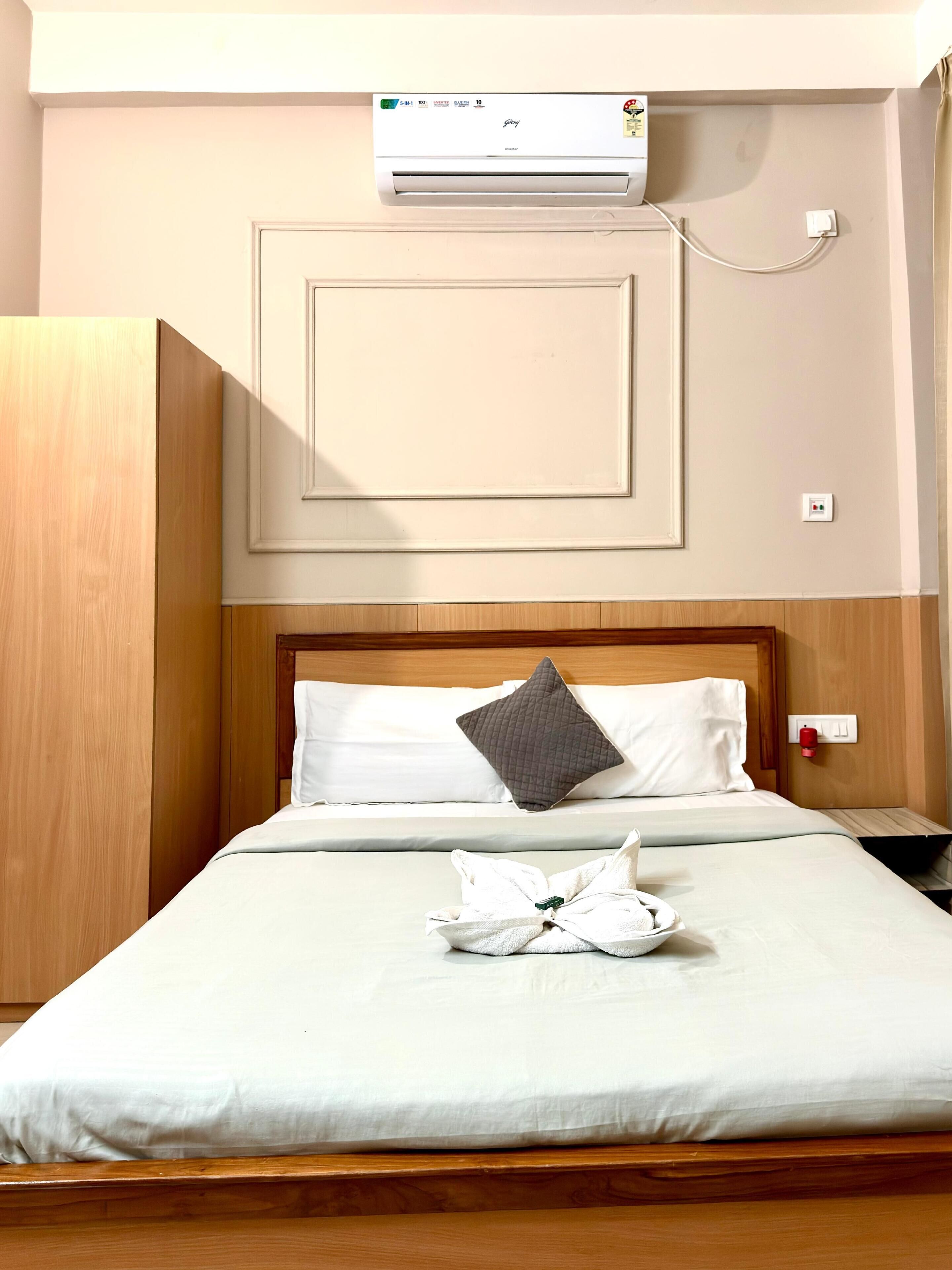 A hotel room with a bed, a bedside table, a wall-mounted air conditioner, and a power outlet.