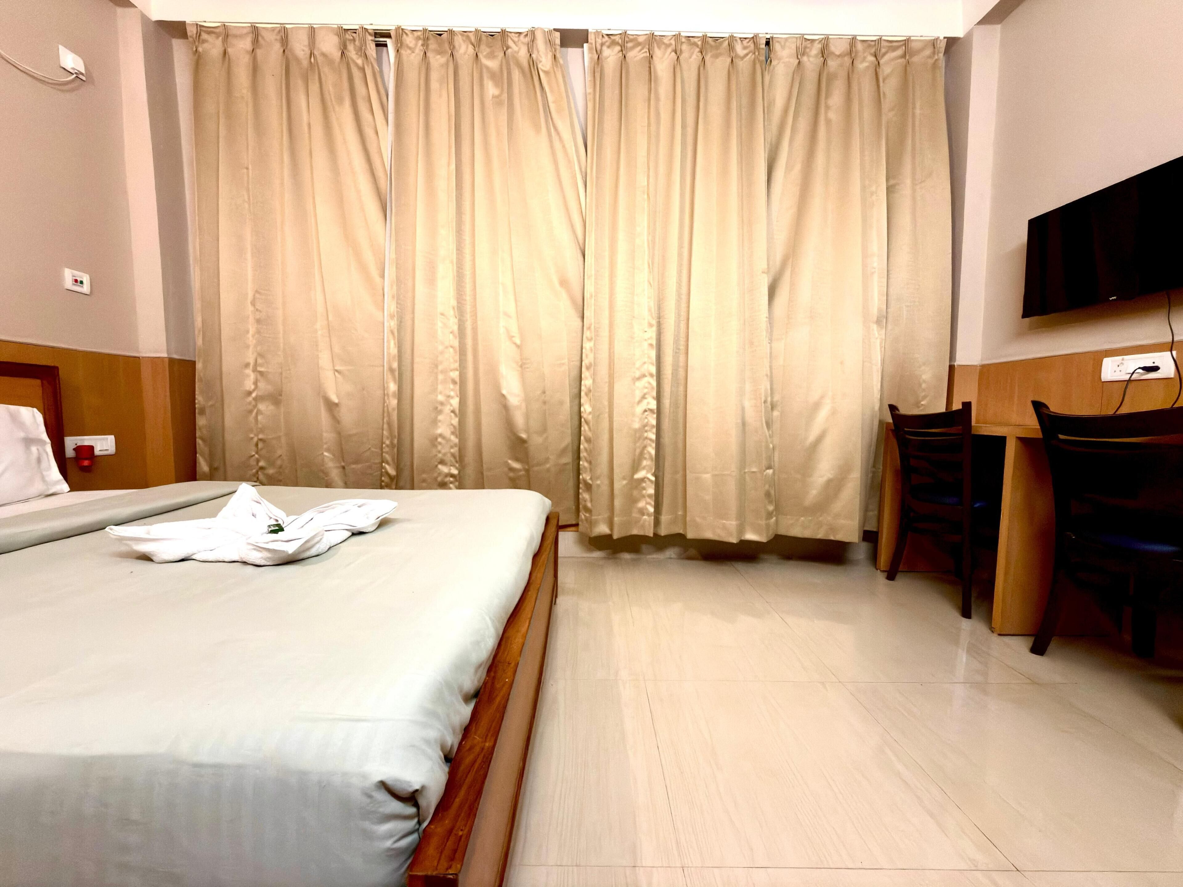 Habitación de hotel con cama, escritorio, silla, televisión y ventana con cortinas.