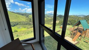 A room with large windows offering a view of a green landscape and a wooden play structure outside. - Loft in the Mantiqueira house. (Gonçalves)
