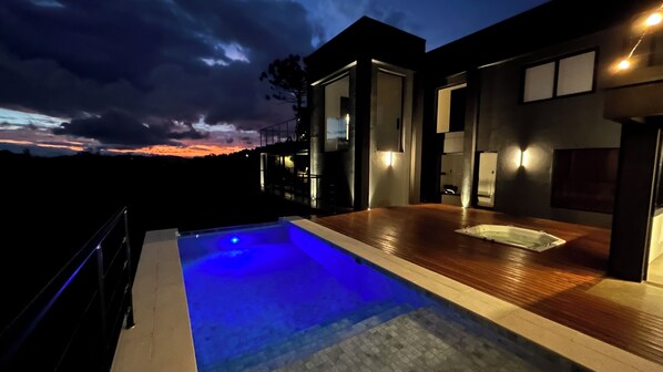 A modern outdoor pool area with a wooden deck and a built-in hot tub, surrounded by a contemporary house with large windows and a view of the sunset.