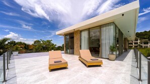 A modern villa with two lounge chairs on a spacious patio, large sliding glass doors, and a view of lush greenery. - Ultra-Modern 6-Bedroom in GolfCourse PH6 (Cha-am)