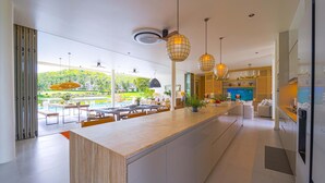 A modern kitchen with a large island, pendant lights, and a view of a pool and greenery. - Ultra-Modern 6-Bedroom in GolfCourse PH6 (Cha-am)