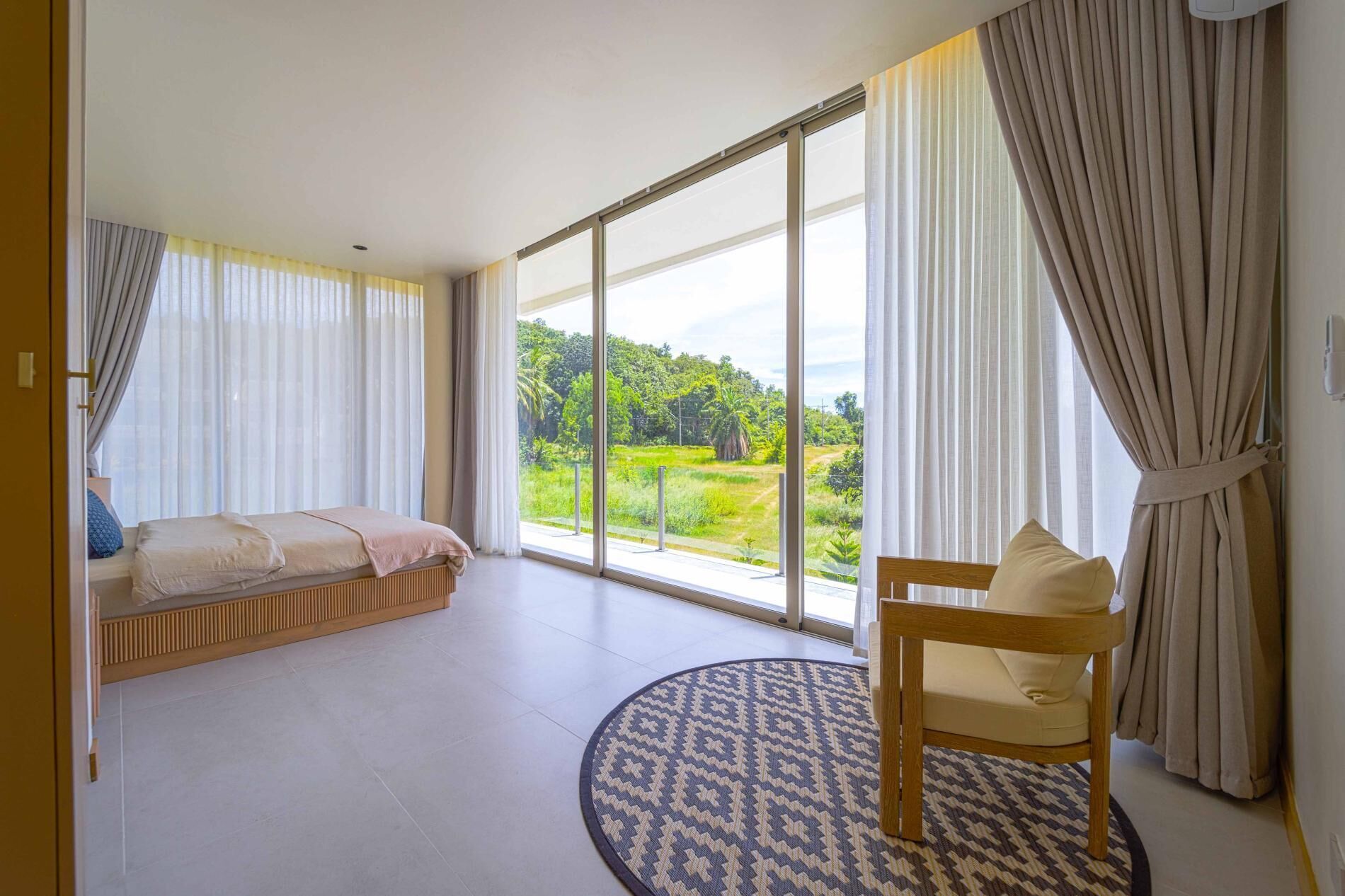 A spacious room with a large window offering a view of greenery outside. There is a bed with a headboard, a chair, and a patterned rug.