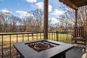 A patio with a built-in fire feature, a wooden chair, and a view of a grassy field and trees.
