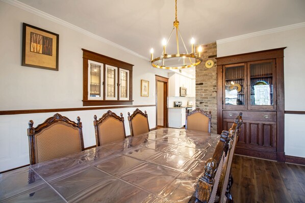 A dining room with a large table covered in plastic, wooden chairs, a chandelier, and a wooden cabinet with glass doors.