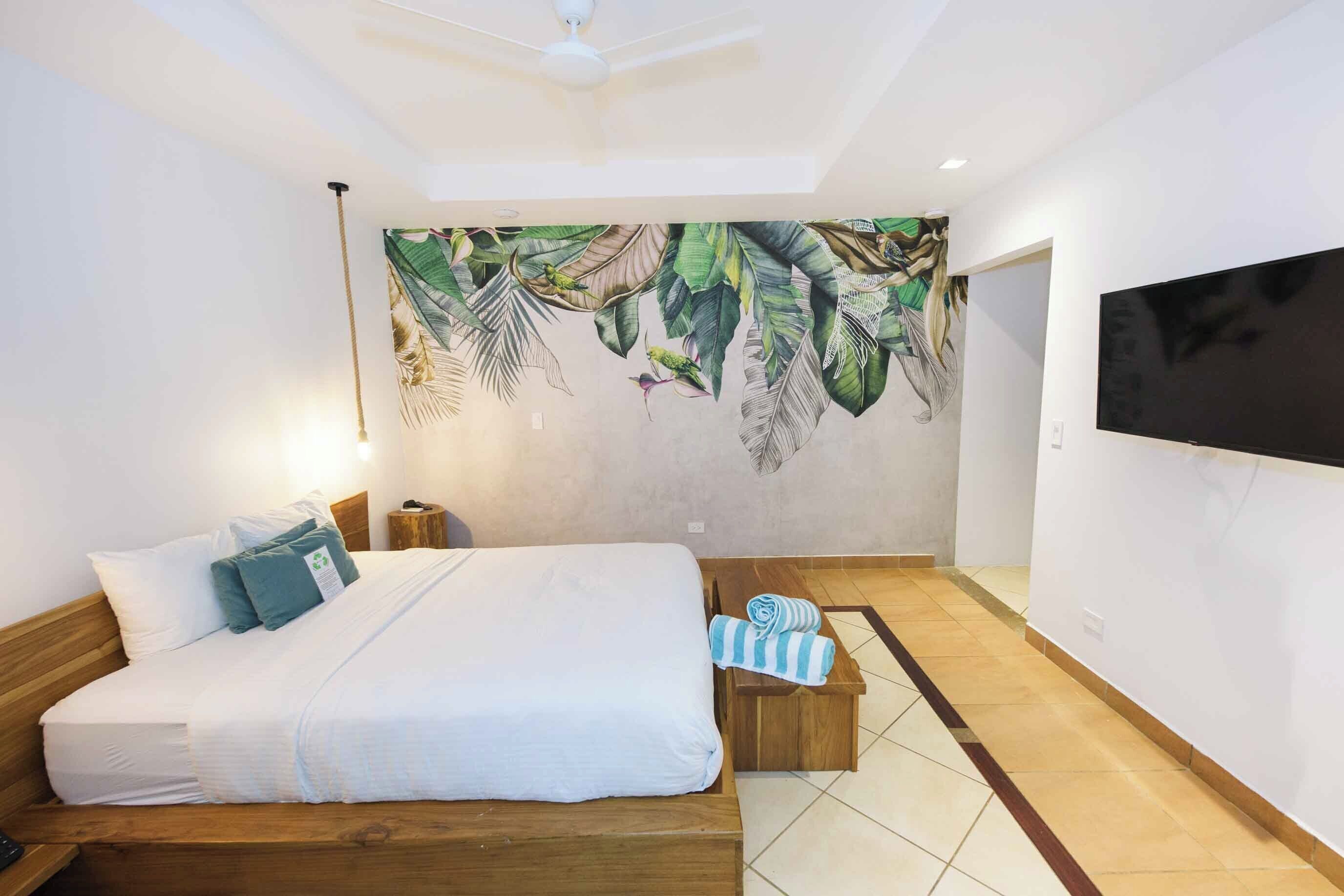 Un dormitorio con cama, televisor montado en la pared, ventilador de techo y un mural de hojas tropicales.