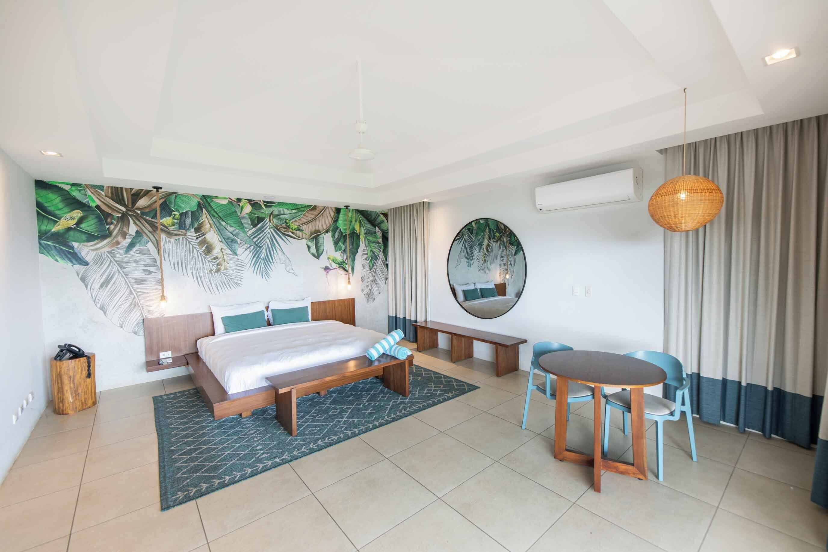 A modern hotel room with a large bed, a dining table with chairs, a round mirror, and a tropical-themed mural on the wall.