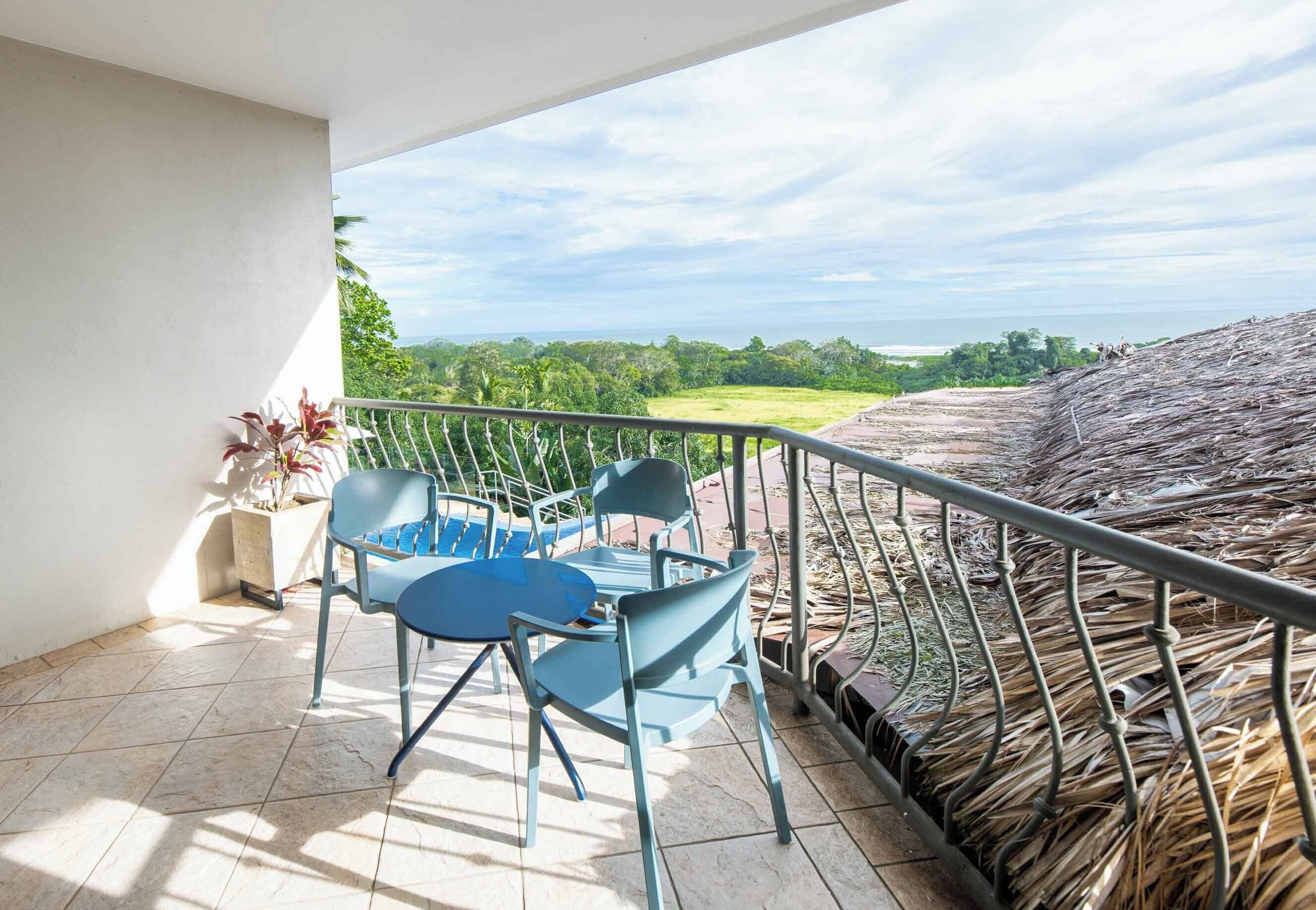 A balcony with a table and chairs, a potted plant, and a view of a lush landscape and ocean.