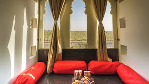 A room with a view of a desert landscape through arched windows, red cushions, and a basket of snacks on a table. - Shreeji Desert Resort (Sam)