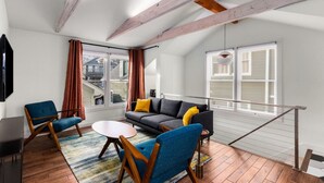 A modern loft-style room with a high ceiling, exposed beams, a gray sofa with yellow cushions, a blue armchair, a small round table, and large windows offering a view of the outside. - Chic ATL Stay | Private, Near BeltLine + WiFi (Atlanta)