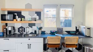 A modern kitchen with a built-in microwave, coffee maker, and kettle, a sink, two wicker chairs, and a window with a view of buildings. - Chic ATL Stay | Private, Near BeltLine + WiFi (Atlanta)