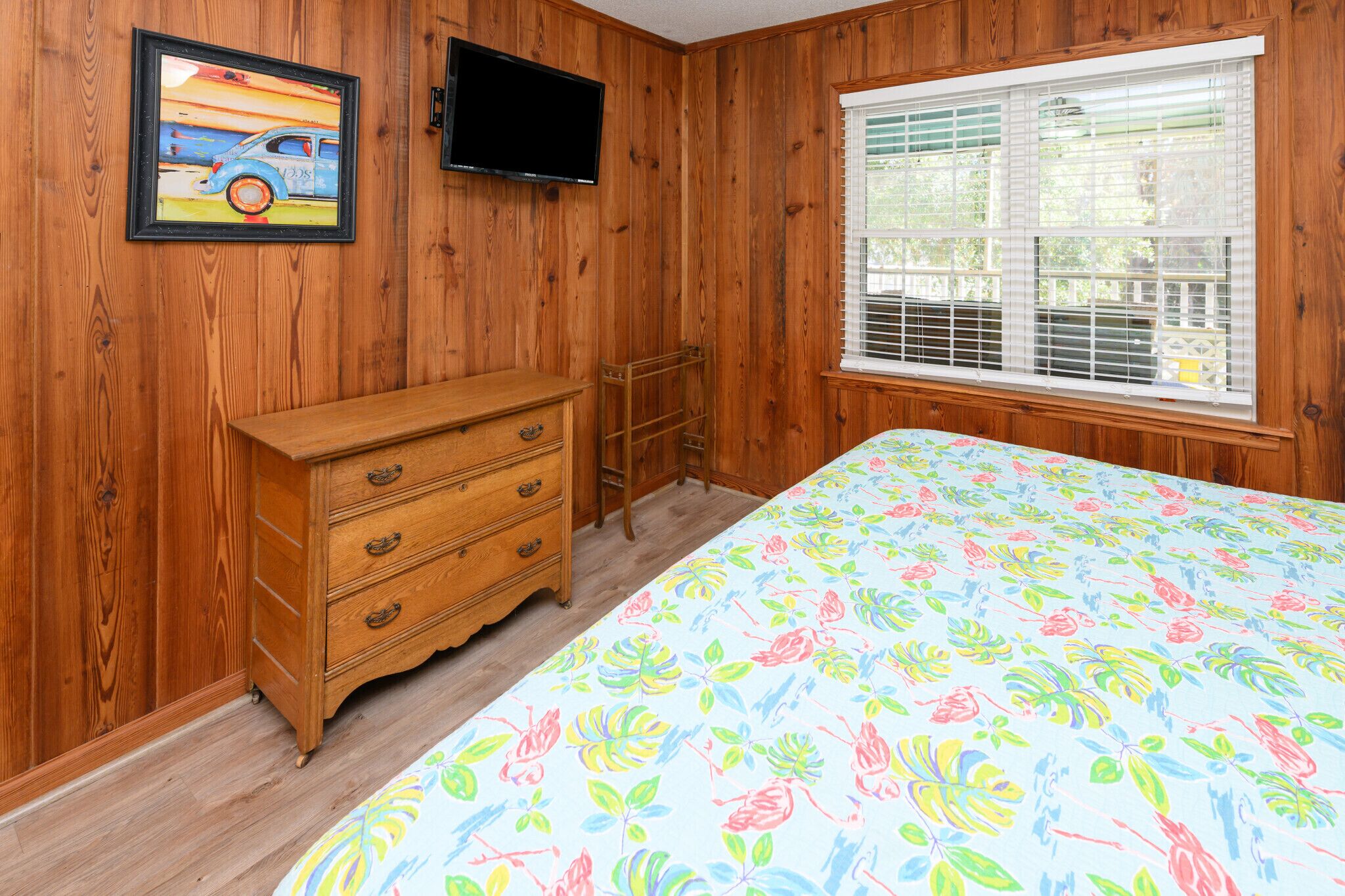 A room with wooden walls, a wooden dresser, a bed with a floral bedspread, a television mounted on the wall, and a window with blinds.