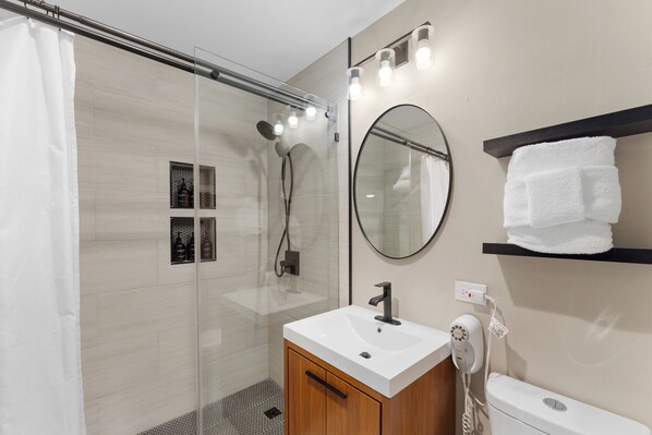 A modern bathroom with a glass shower enclosure, a white sink, a round mirror, and a towel rack. - Slope-Side Luxury Condo - Pool with Slide, Hot Tubs, & More! (Keystone)