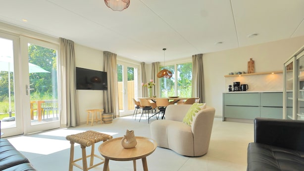 A modern living room with a sofa, a round wooden table, and a beige armchair. There is a dining area with a table and chairs, and a kitchen area with cabinets and a countertop. - Modern vakantiehuis aan de kust! (Callantsoog)