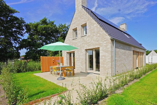 Une maison moderne avec un toit en pente, une façade en briques et un patio agrémenté d’une table et de chaises sous un parasol vert.