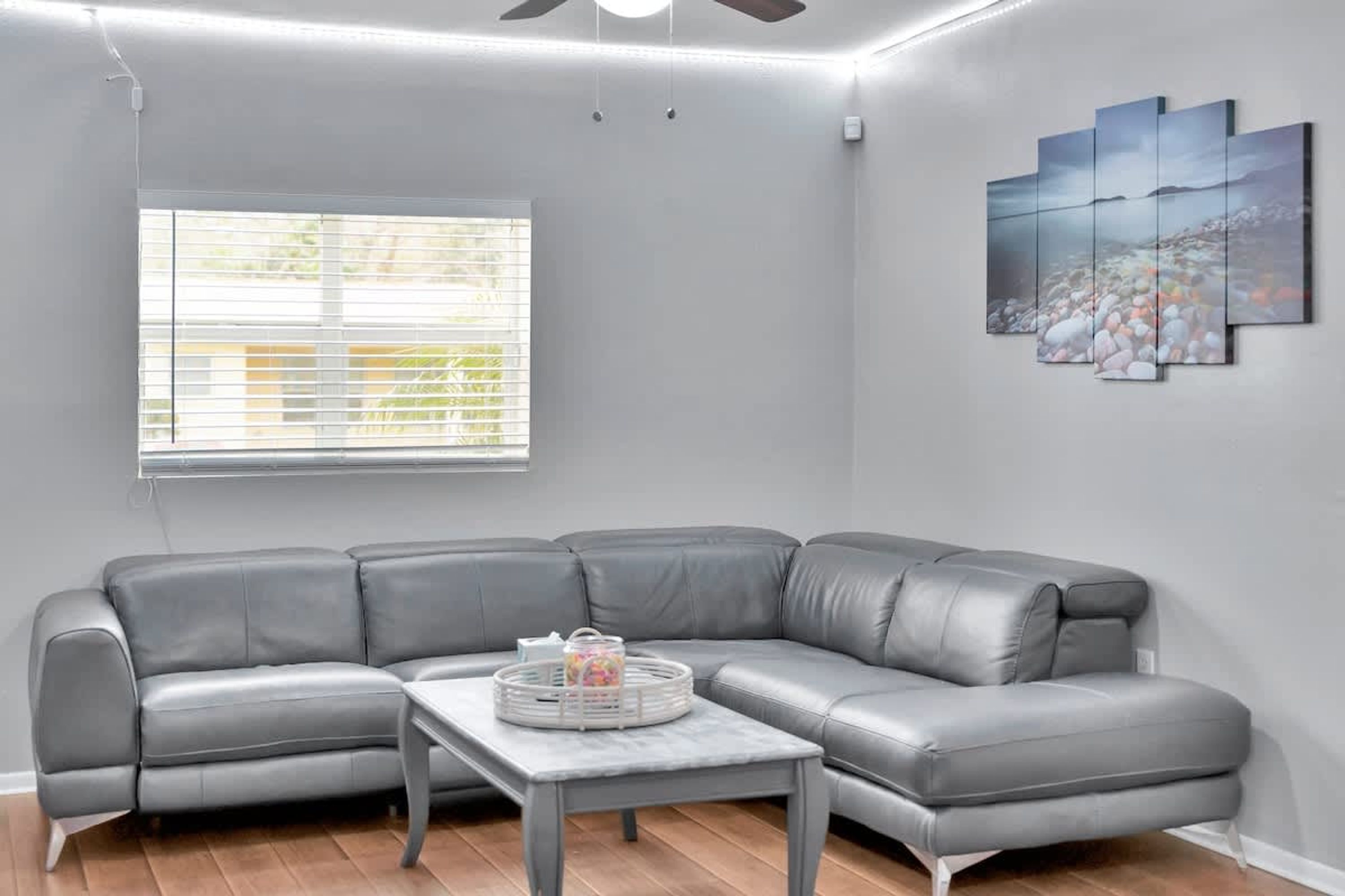 A modern living room with a gray leather sectional sofa, a small table with a basket, a window with blinds, and a wall-mounted art piece.