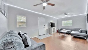 A spacious living room with a grey sofa, a coffee table, and a TV on a white cabinet. There are two ceiling fans and a door leading to another room. - Family Pet Friendly Getaway With Porch Fire Pit Yard (Tampa)