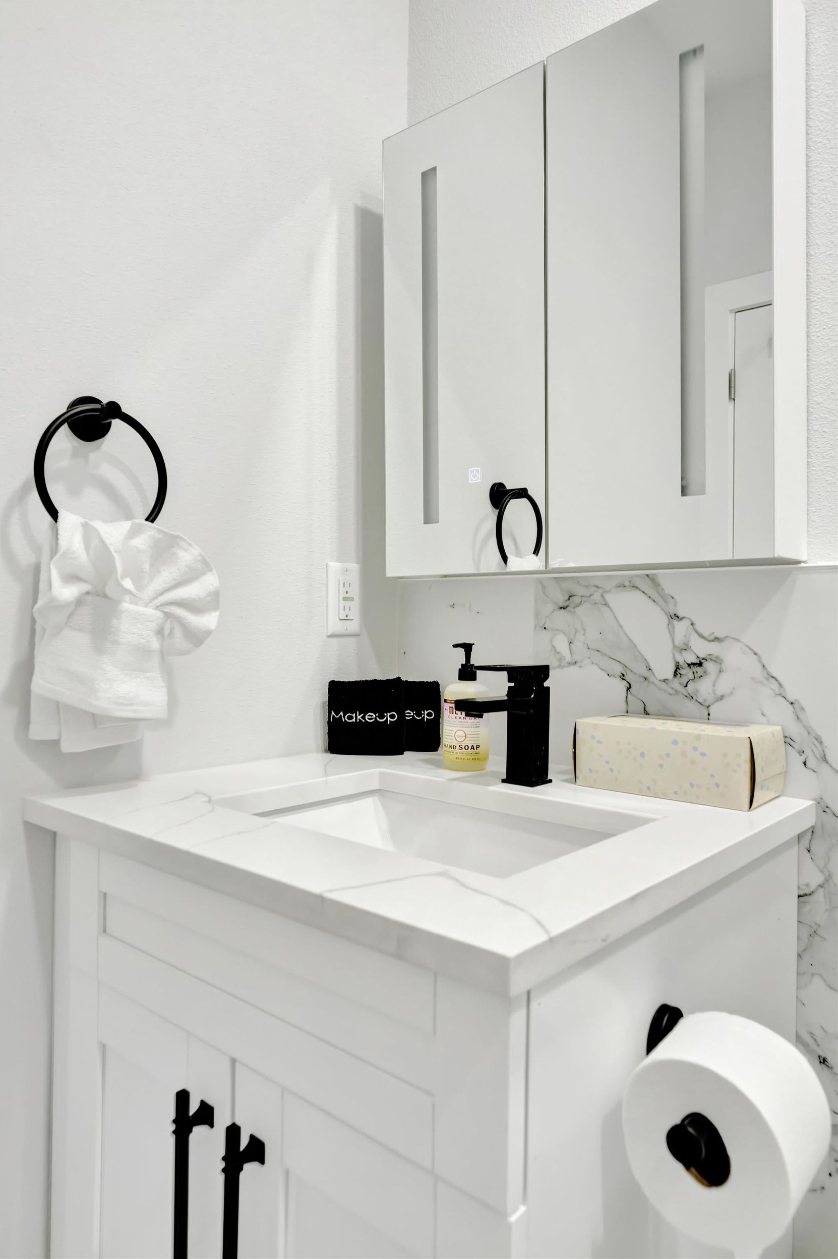 Une salle de bain moderne avec un meuble-lavabo blanc, un miroir, un porte-serviettes, une trousse de maquillage, un distributeur de savon et un porte-rouleau de papier toilette.