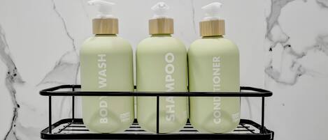 Three bottles of body wash, shampoo, and conditioner on a black metal shelf against a white marble wall.