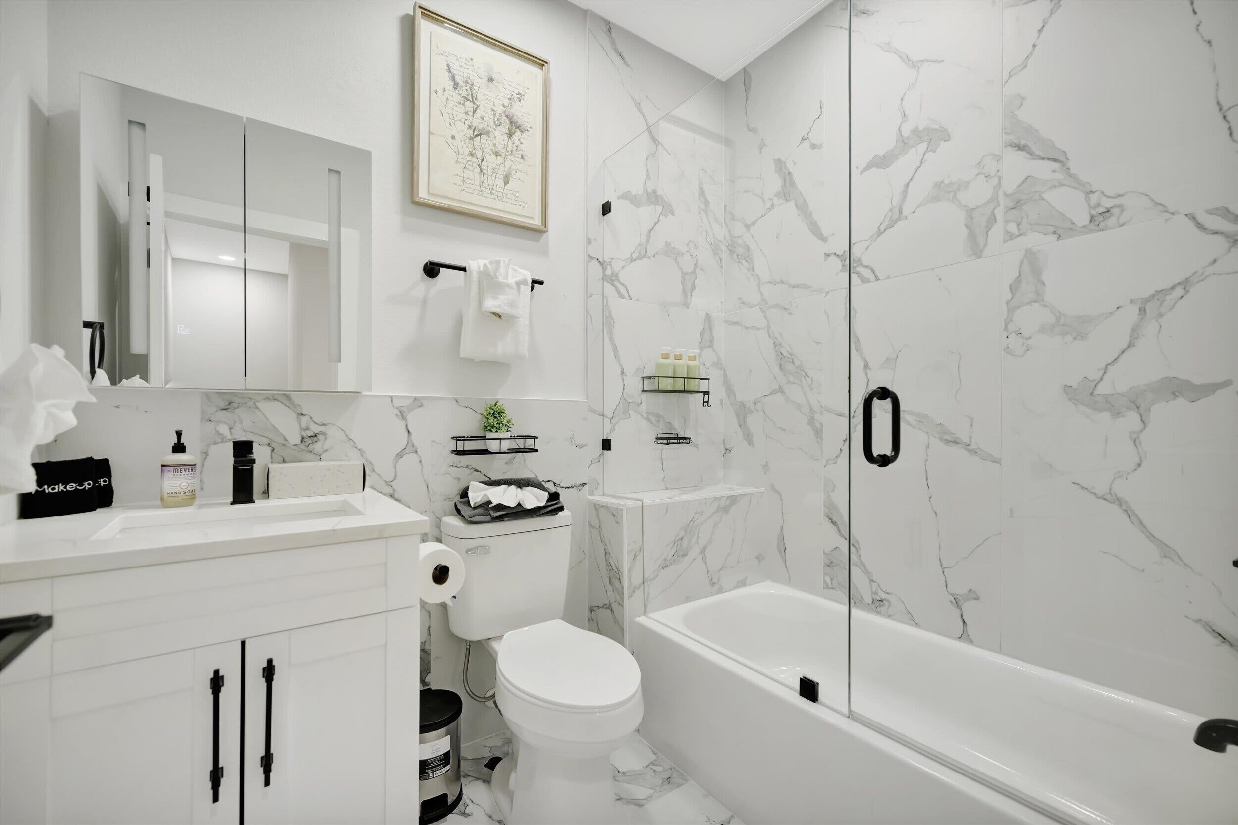 A modern bathroom with a white marble wall, a white toilet, a white bathtub, and a white sink with a mirror above it.