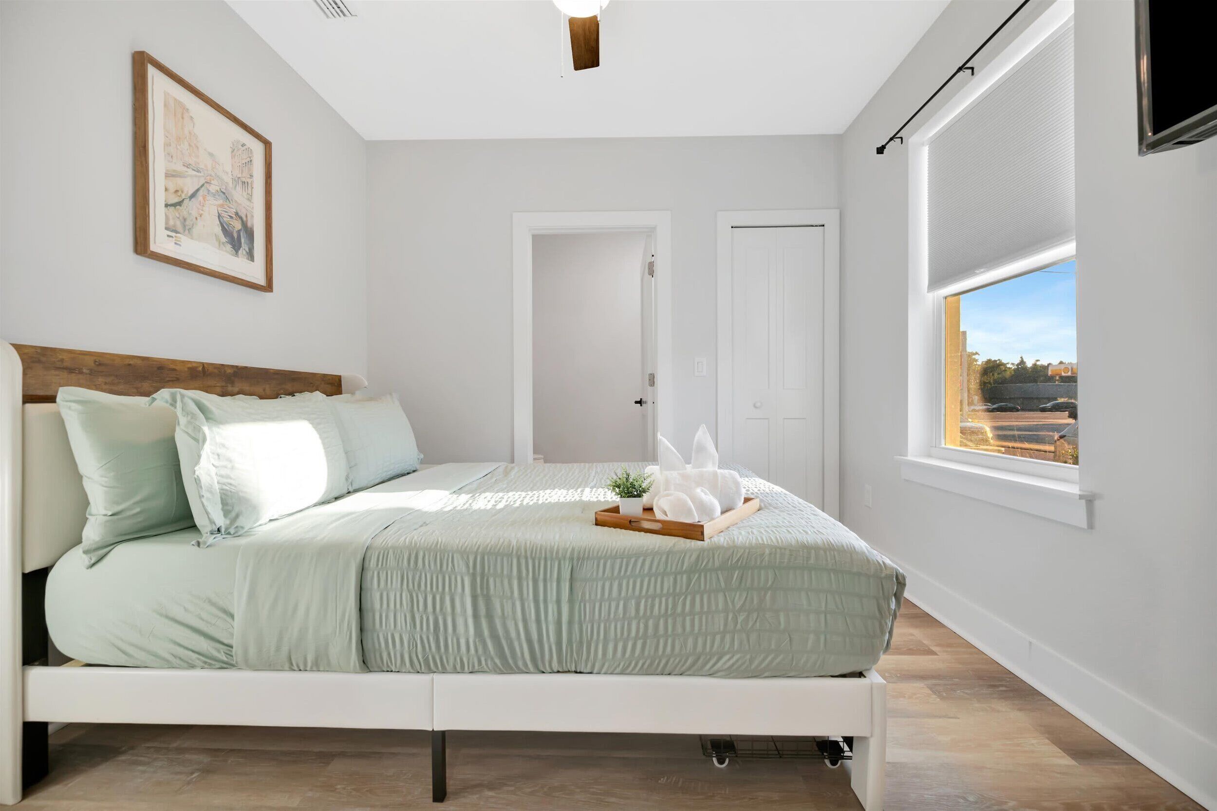 Un dormitorio con cama, ventana con vistas, puerta y un cuadro enmarcado en la pared.