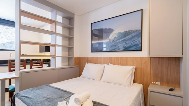 A hotel room with a bed, a desk, a chair, a TV, and a large ocean wave painting on the wall. - Studio 290 m From Praia Mole Vds0005 (Florianópolis)