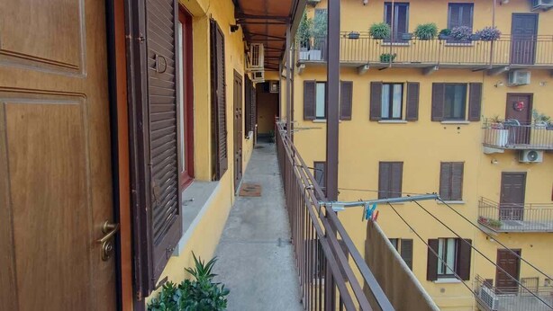 Un balcone stretto con una porta e persiane di legno, affacciato su un edificio giallo con molte finestre e balconi.
