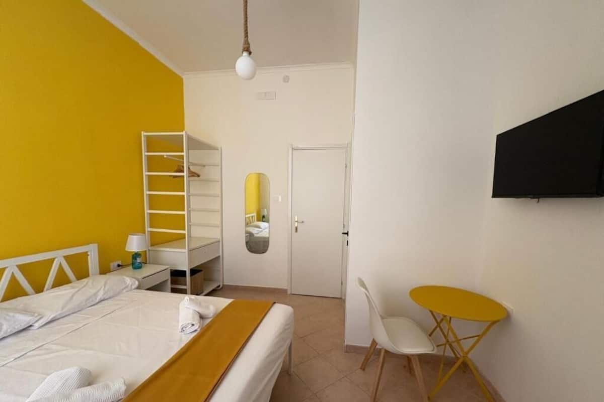 A bedroom with a bed, a white wardrobe, a small table with a chair, a mirror, and a TV mounted on the wall.