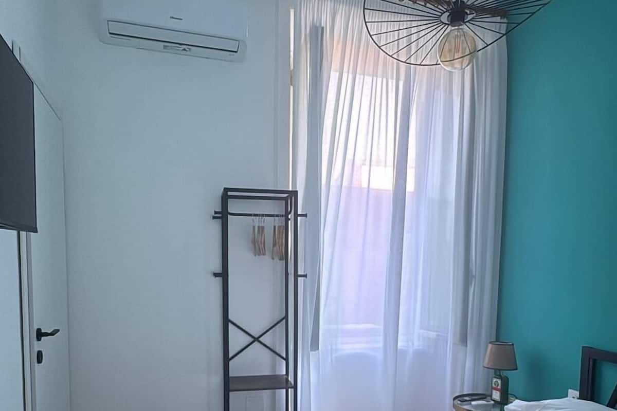A hotel room with a bed, a wardrobe, a mirror, a hanging light fixture, and a window with sheer curtains.