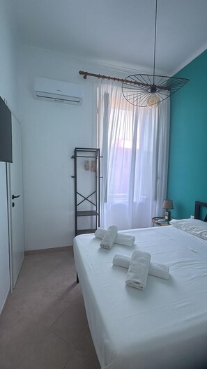 Une chambre d’hôtel comprenant un lit, une armoire, un miroir, un luminaire suspendu et une fenêtre avec des rideaux légers.