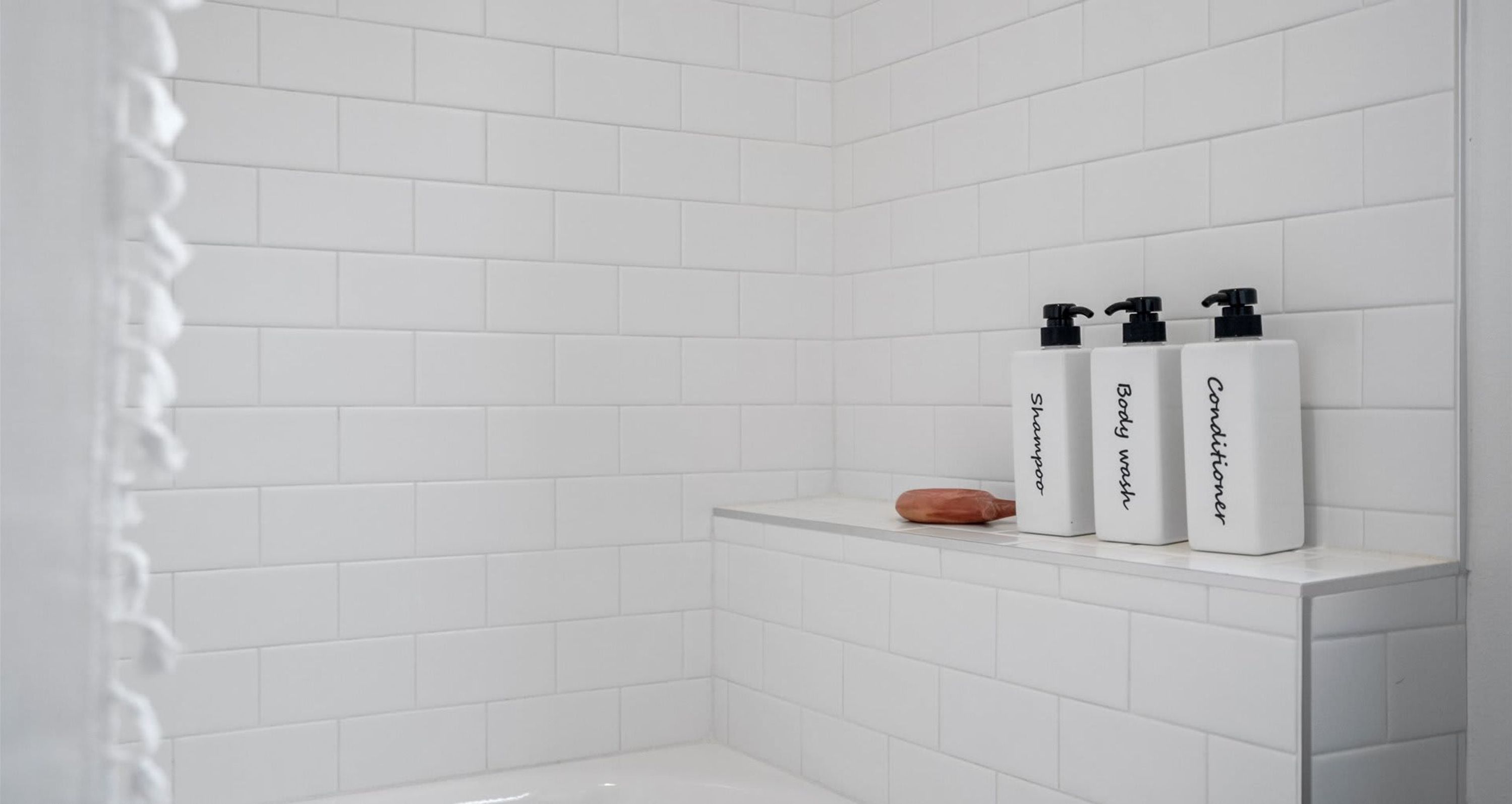 Ein Badezimmer mit weißen Fliesenwänden und einem Regal mit drei Flaschen, beschriftet mit 'Body wash', 'Shampoo' und 'Conditioner'.