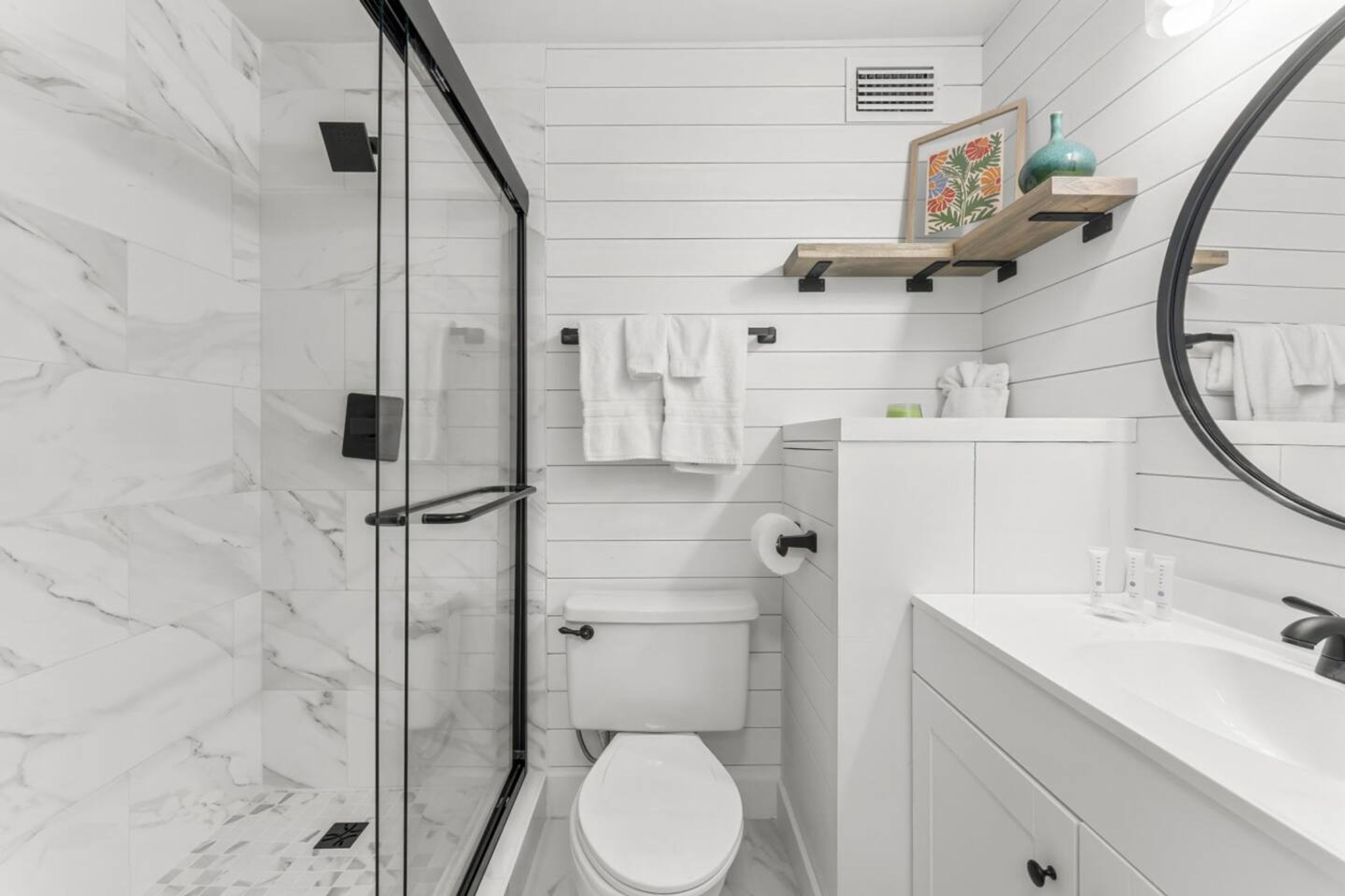 Een moderne badkamer met een glazen douchecabine, witte marmeren wanden, een wit toilet en een wastafel met een spiegel en planken.