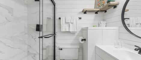 Een moderne badkamer met een glazen douchecabine, witte marmeren wanden, een wit toilet en een wastafel met een spiegel en planken.