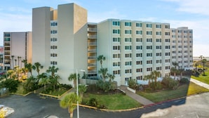 A multi-story hotel with a modern design, featuring a central courtyard with palm trees and a parking area in front. - Sea the Gulf - Top of the Gulf 809 - Luxury Resort (Panama City Beach)
