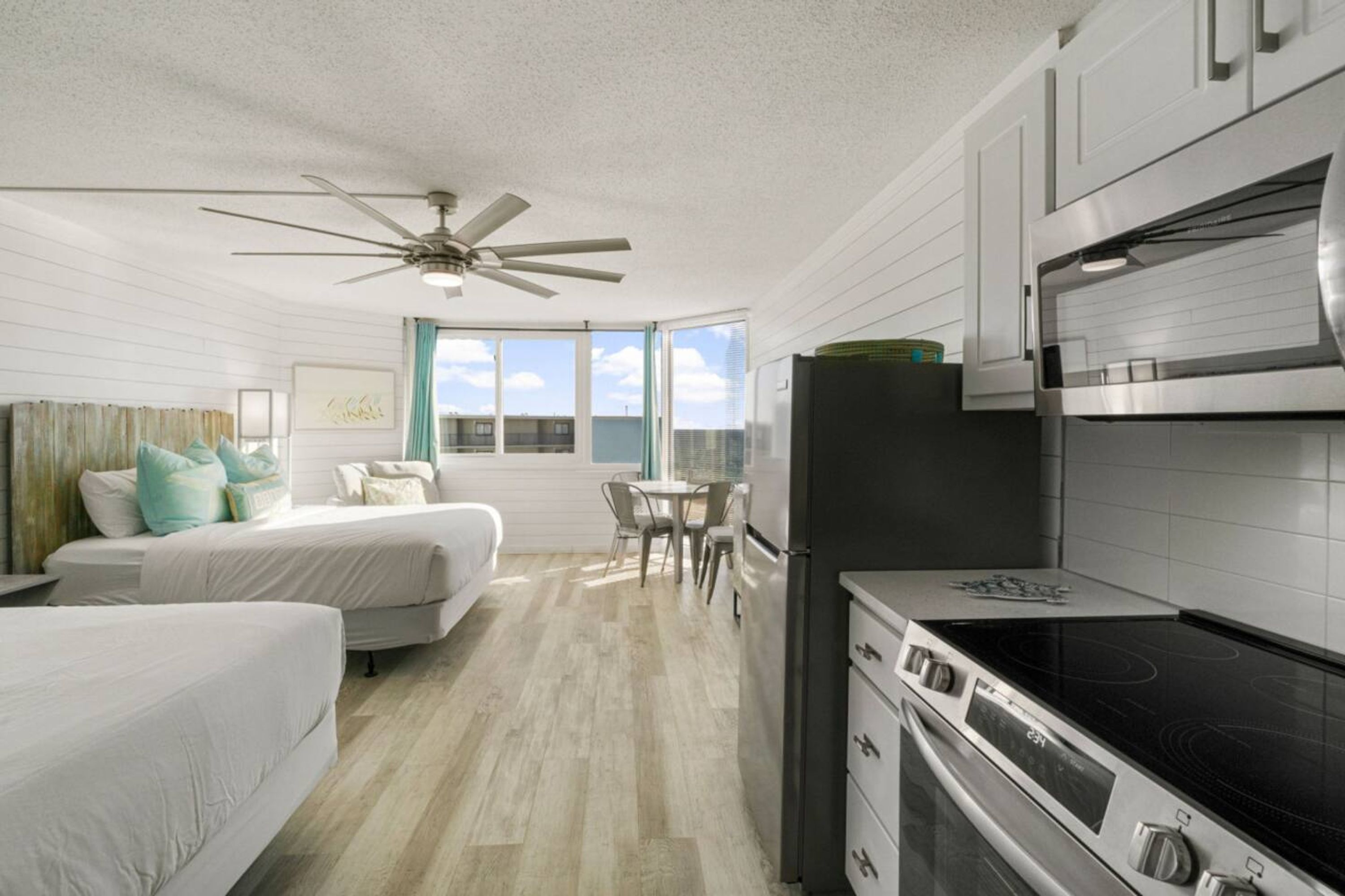 Habitación de hotel compacta con kitchenette, dos camas, zona de comedor y un ventanal con vistas a la ciudad.