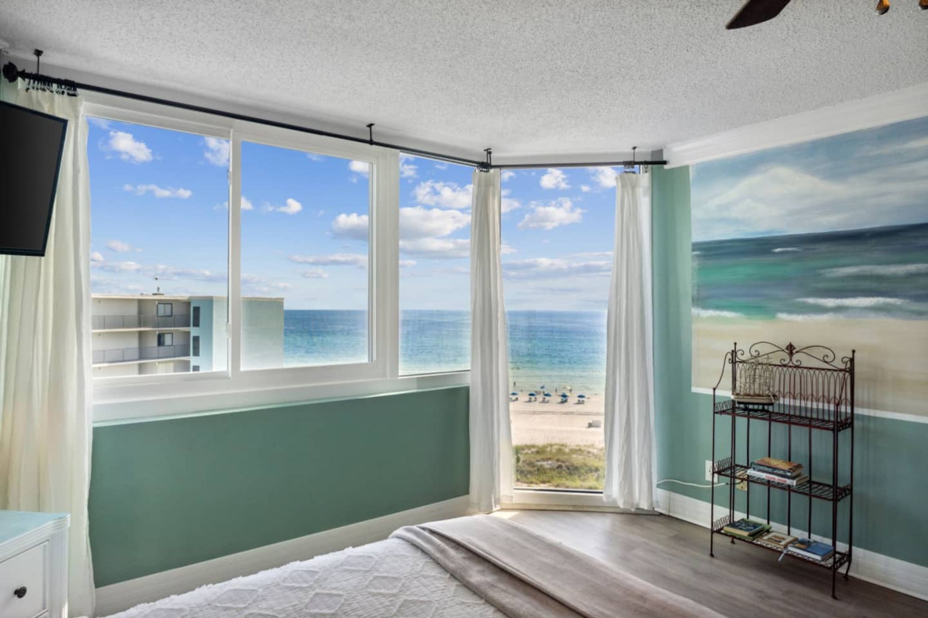 A bedroom with a large window overlooking a beach, a bookshelf, and a painting of the ocean.