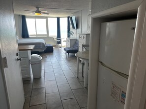 A compact hotel room with a bed, a small table, a chair, a refrigerator, and a trash bin.