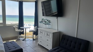 Hotellihuone, jossa on sänky, televisio, lipasto, tuoli, pieni pöytä ja näkymä rannalle.