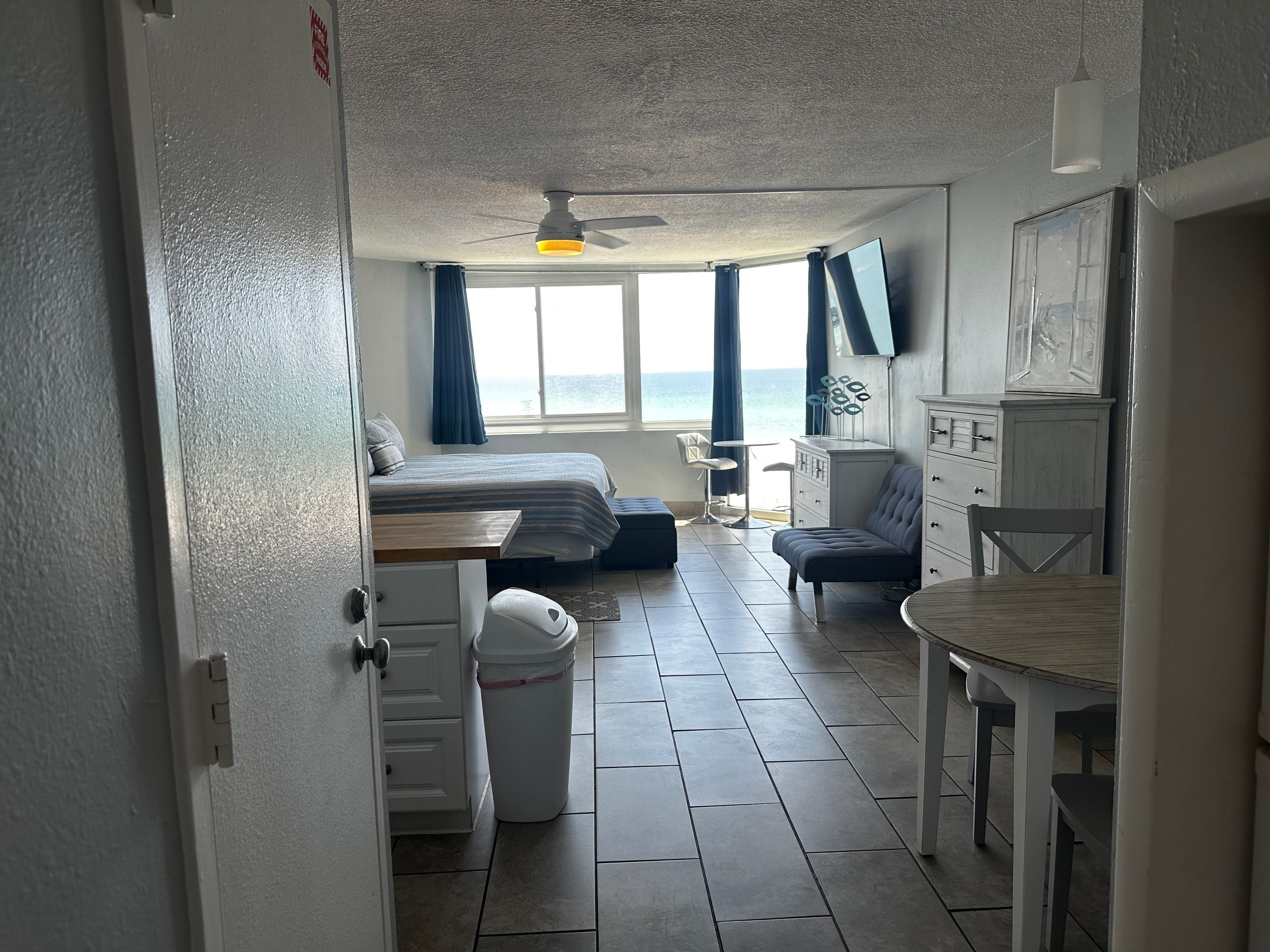 Habitación pequeña con cama, una mesita, una silla y vista al mar a través de una ventana.
