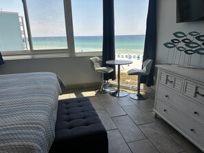 A hotel room with a bed, a small table with two chairs, a dresser, and a view of the beach.