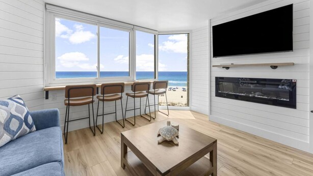 A room with a large window overlooking a beach, a flat-screen TV mounted on the wall, and a small table with a turtle decoration. - Beach Front Ocean View Condo - Top of the Gulf 521 (Panama City Beach)