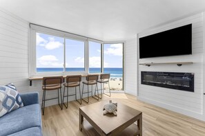 Ein Zimmer mit großem Fenster mit Blick auf einen Strand, einem an der Wand befestigten Flachbildfernseher und einem kleinen Tisch mit einer Schildkrötenfigur.