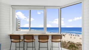 A room with a large window overlooking a beach, featuring four bar stools and a wooden table. - Beach Front Ocean View Condo - Top of the Gulf 521 (Panama City Beach)