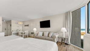 Ein Hotelzimmer mit einem großen Bett, einer Couch, einem Schreibtisch mit Stuhl, einem Fernseher und Blick auf den Strand.
