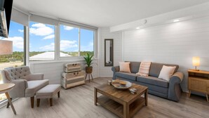 A modern living room with a sofa, armchair, coffee table, and a view of the outdoors through large windows. - Top of the Gulf 622 - Beach Front Condo Resort (Panama City Beach)