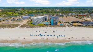Vista aérea de um resort à beira-mar, com um edifício grande, várias piscinas e uma praia de areia com guarda-sóis.