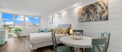 A modern bedroom with a large window, a bed, a bedside table, and a dining area with a table and chairs.